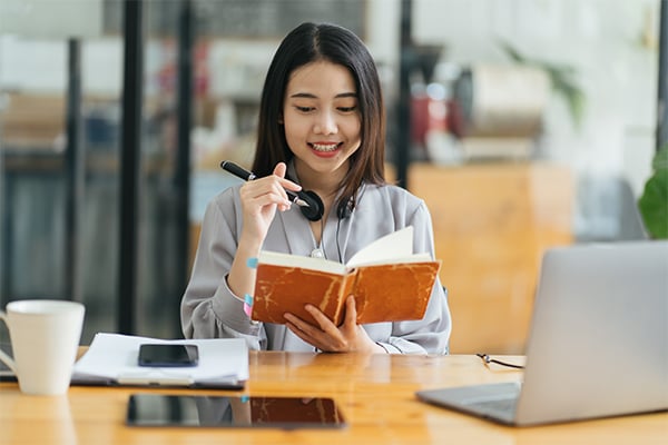 Image of a girl studying in front of a laptop with a book open that she is writing in 